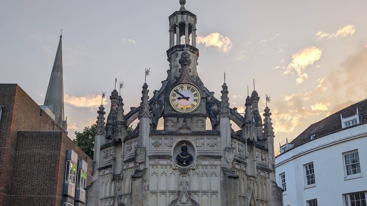 The carved stone Market Cross in Chichester city centre at sunset, with its clock and the cathedral spire behind