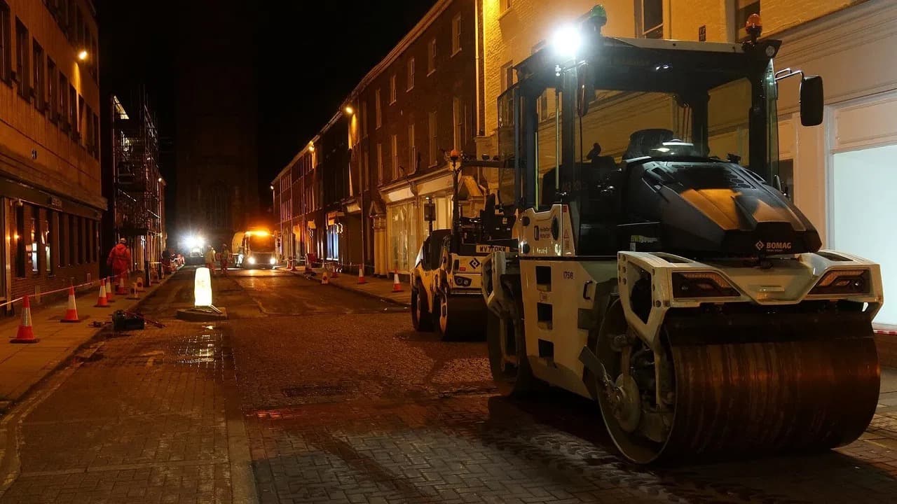 Night road resurfacing on a town street, with two asphalt rollers beside traffic cones and workers in high visibility clothing