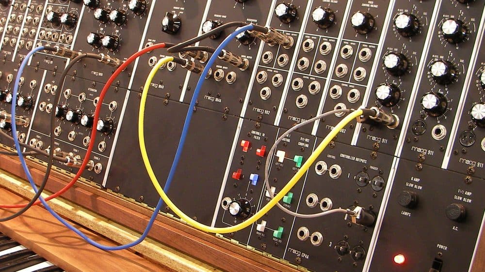 A close view of a modular synthesizer, with coloured patch cables looping across rows of black control panels, knobs and sockets above a wooden keyboard