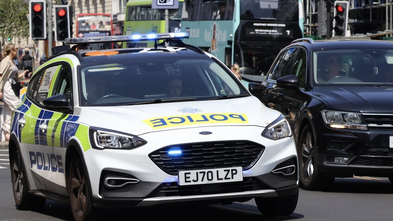 A white Sussex Police patrol car with blue lights on driving along a busy city street with a bus behind it