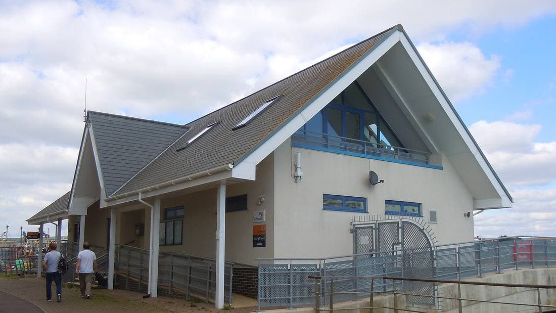 The pitched-roof RNLI Newhaven lifeboat station building on a sunny day with clouds, with two people walking along the quay path beside it