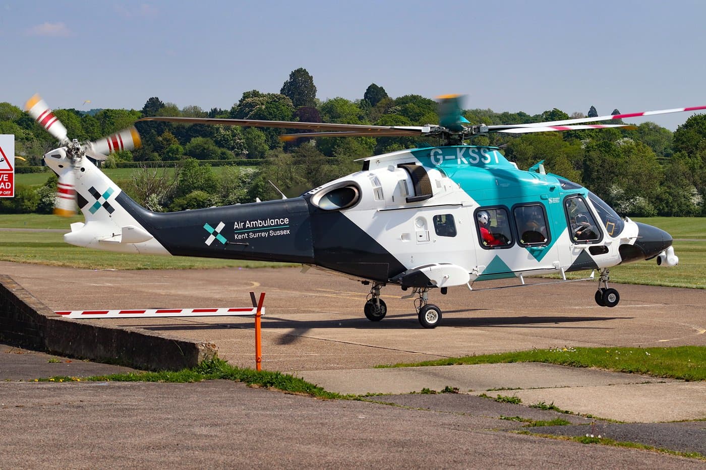 A white and teal Kent Surrey Sussex air ambulance helicopter parked on an airfield with its rotor blades turning