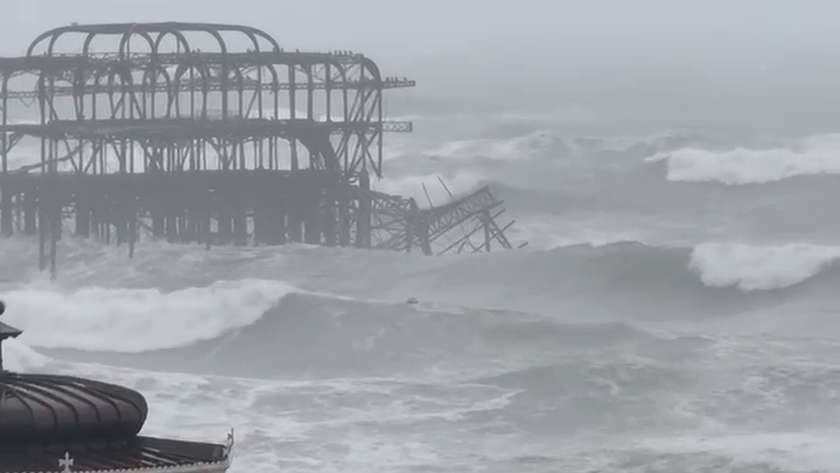 The skeletal iron frame of the West Pier standing in rough grey sea with a fallen section of girders tilted in the water beside it, and the roof of the seafront bandstand in the foreground