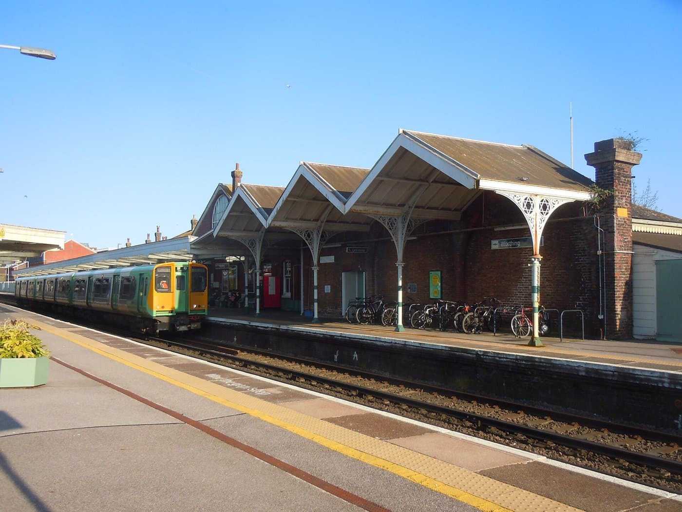 A green and yellow passenger train standing at a platform with a covered brick station building and bicycles beside it