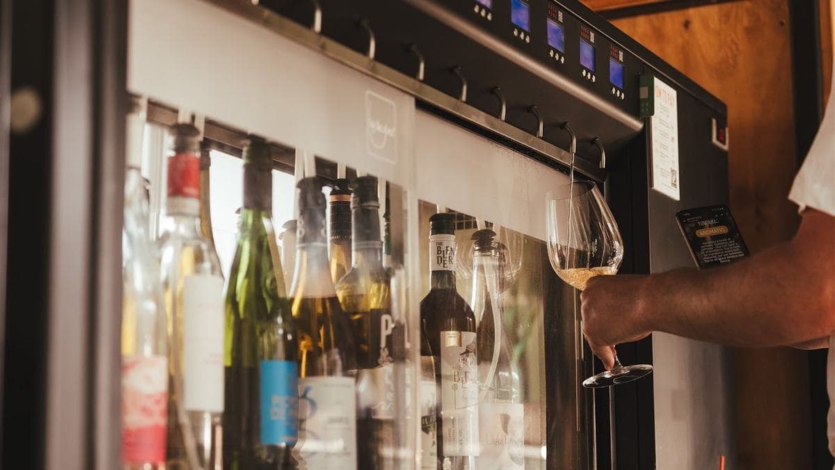 A customer pouring a glass of white wine from a self-pour wine machine at a Vagabond bar, with rows of bottles behind