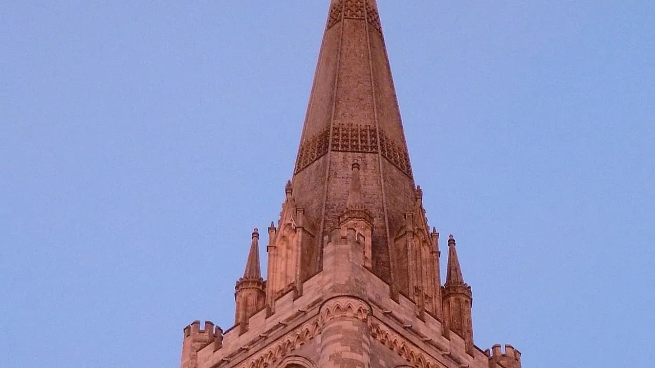 The spire and tower of Chichester Cathedral lit pink by the setting sun against a blue sky