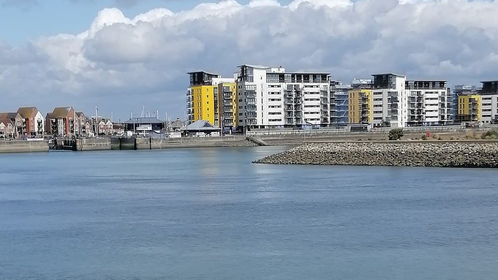 Calm water at Sovereign Harbour in Eastbourne with blocks of flats on the far shore, a stone breakwater and a shingle bank in the foreground