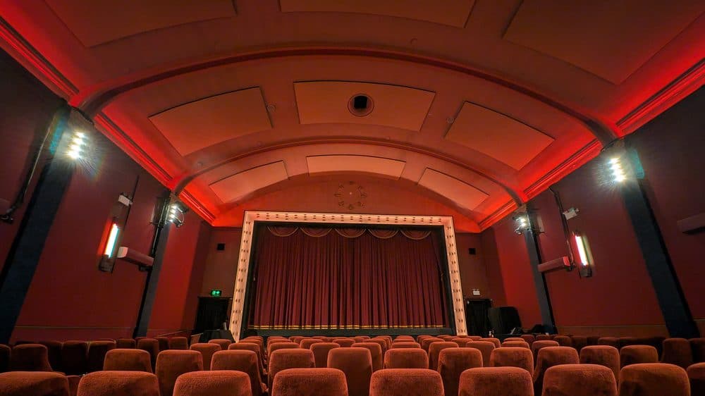 The empty auditorium of a Brighton cinema, with rows of red velvet seats facing a curtained screen under a curved, red-lit ceiling
