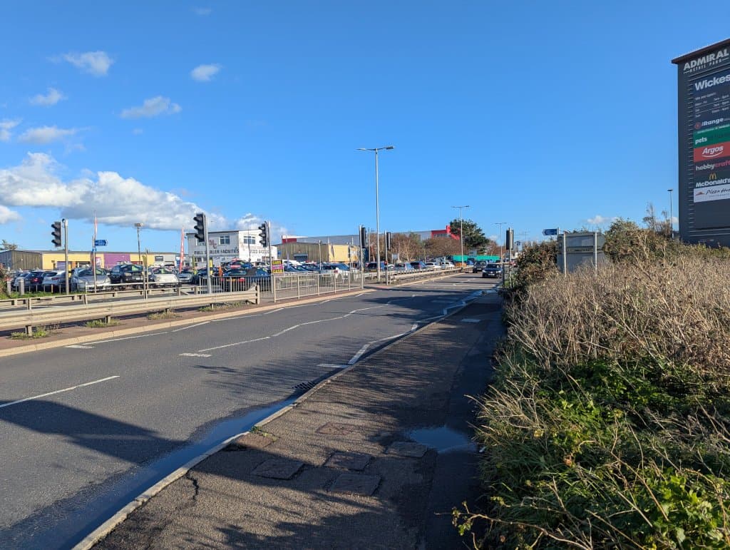 A busy dual carriageway with traffic lights and a retail park car park beside it on a sunny day
