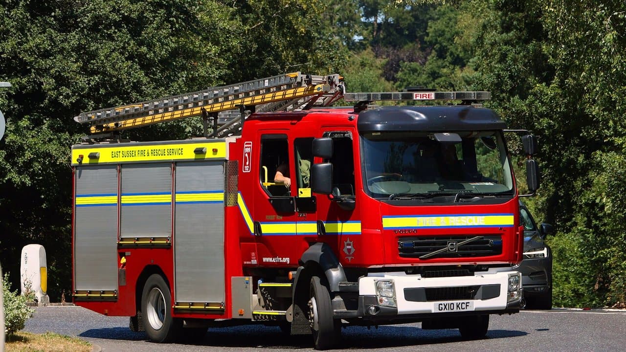 An East Sussex Fire and Rescue Service fire engine with a ladder on its roof turning a corner on a tree-lined road