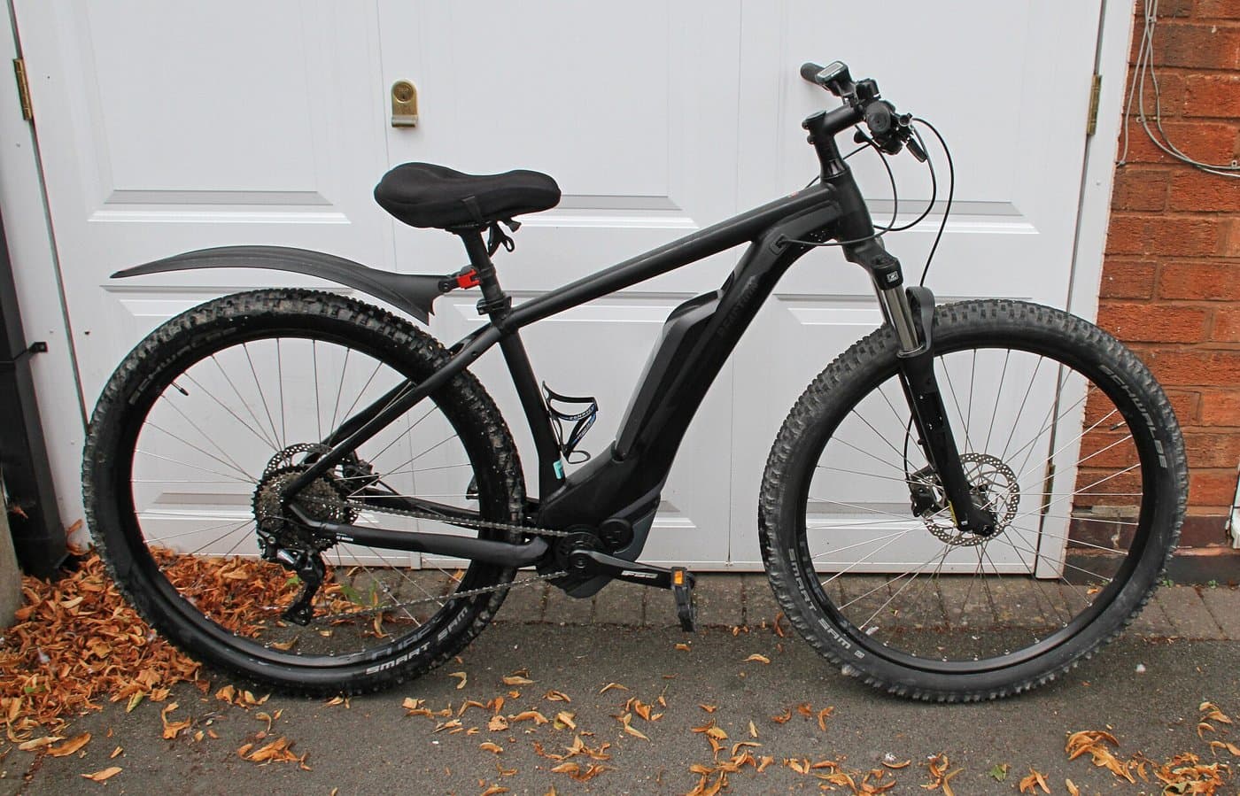 A black electric mountain bike with a mudguard and a battery in the frame, standing on a paved drive in front of a white door