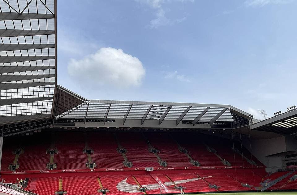 The red seats and pitch inside Anfield, Liverpool’s stadium, seen from the corner of the Anfield Road stand under a cloudy blue sky
