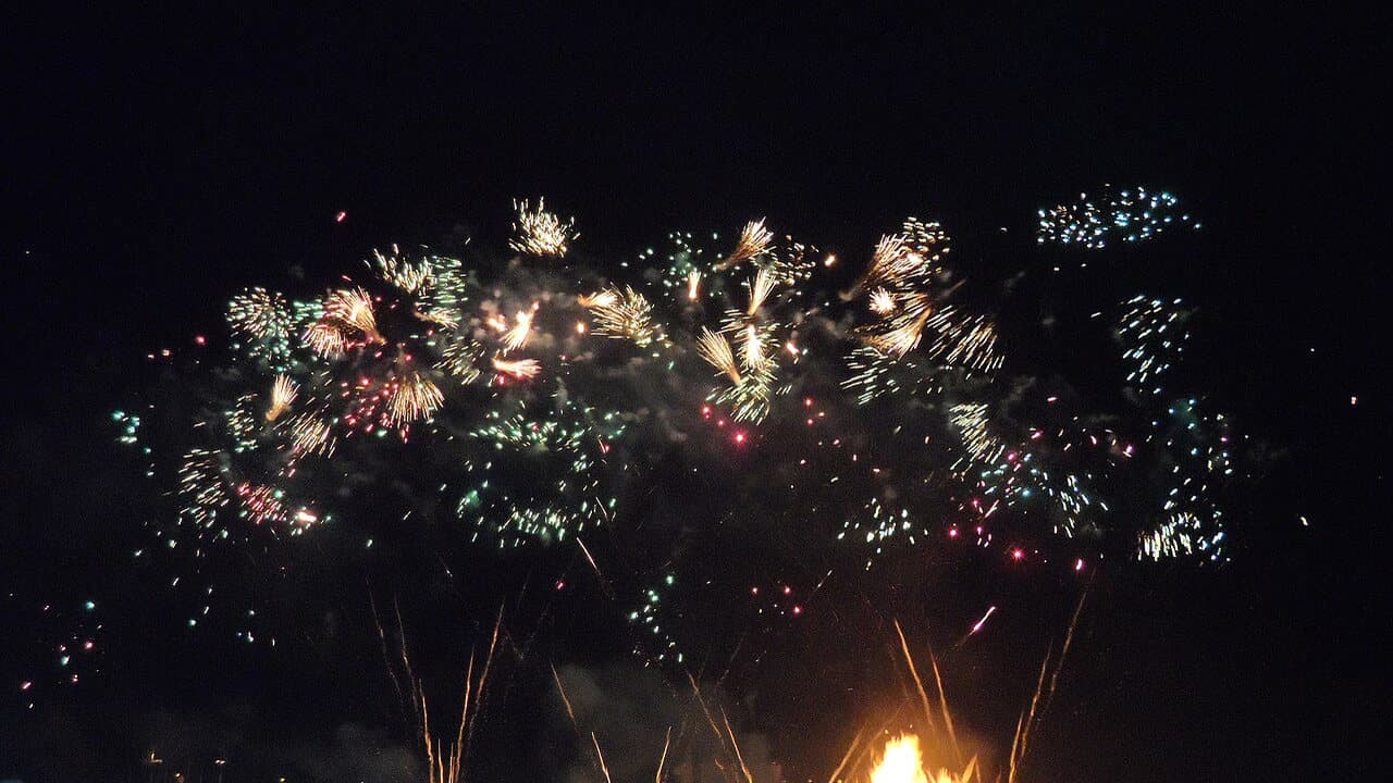 Fireworks bursting in white, gold and red over a dark night sky with a bonfire glowing at the bottom of the picture