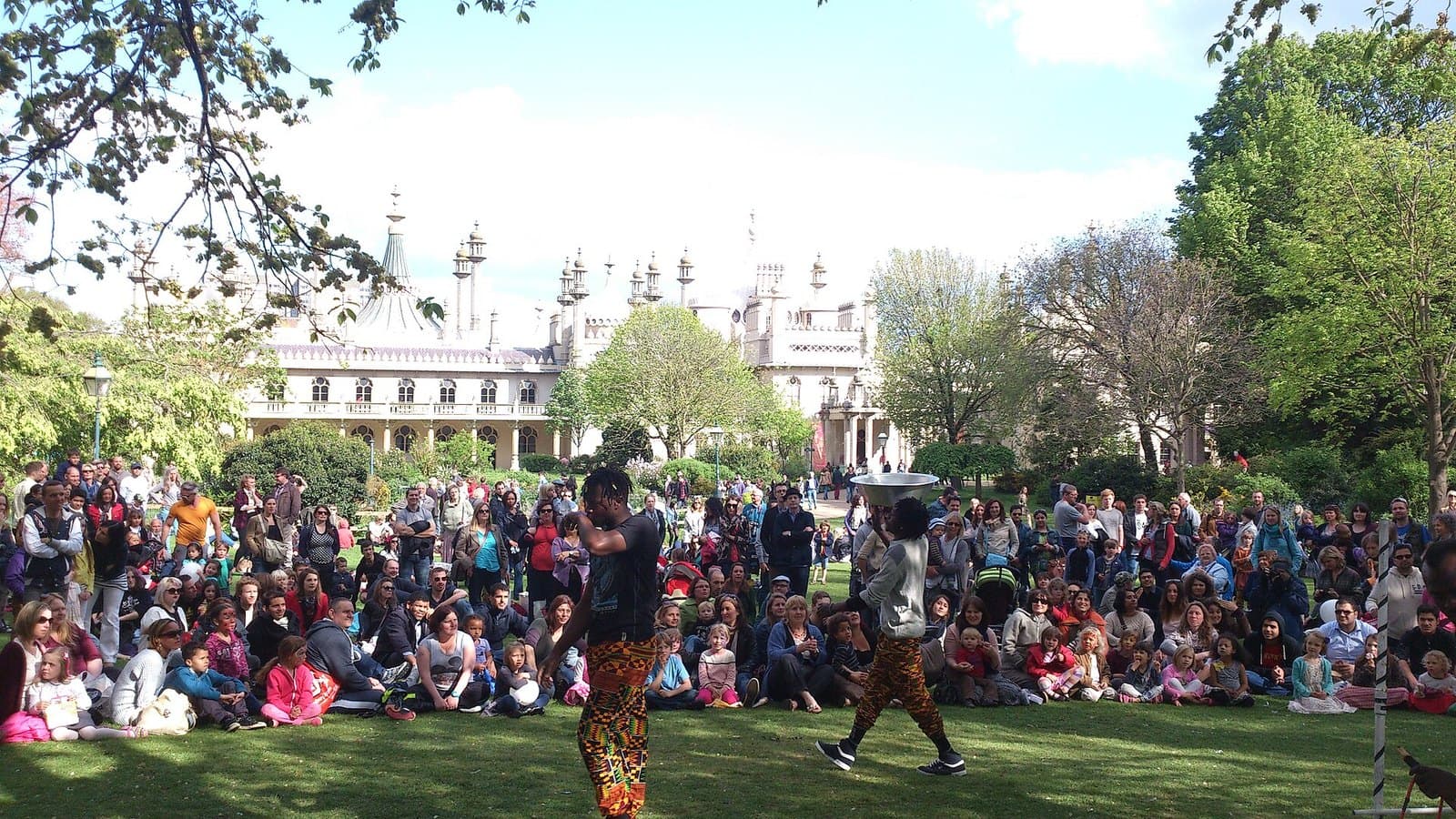 Performers in front of a seated crowd in the Pavilion Gardens, with the Royal Pavilion behind, during Fringe City at Brighton Fringe