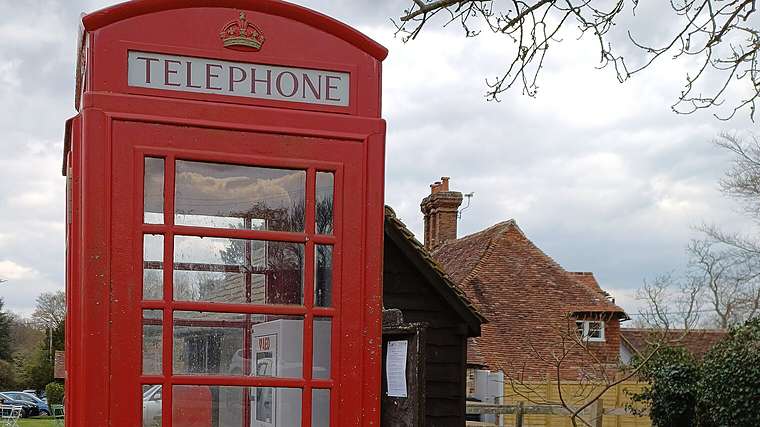 A red K6 telephone box beside a village green in Lurgashall, West Sussex, with a tiled roof house behind it