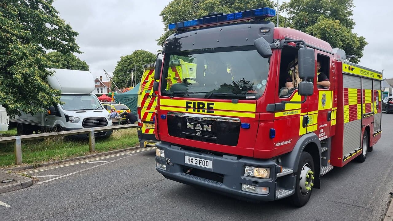 A red and yellow West Sussex Fire and Rescue Service fire engine with its blue lights on, driving along a road