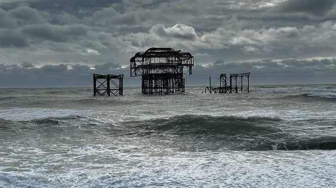 The West Pier standing in rough grey sea as three separate pieces of ironwork under heavy cloud, with waves breaking on the shingle beach in the foreground