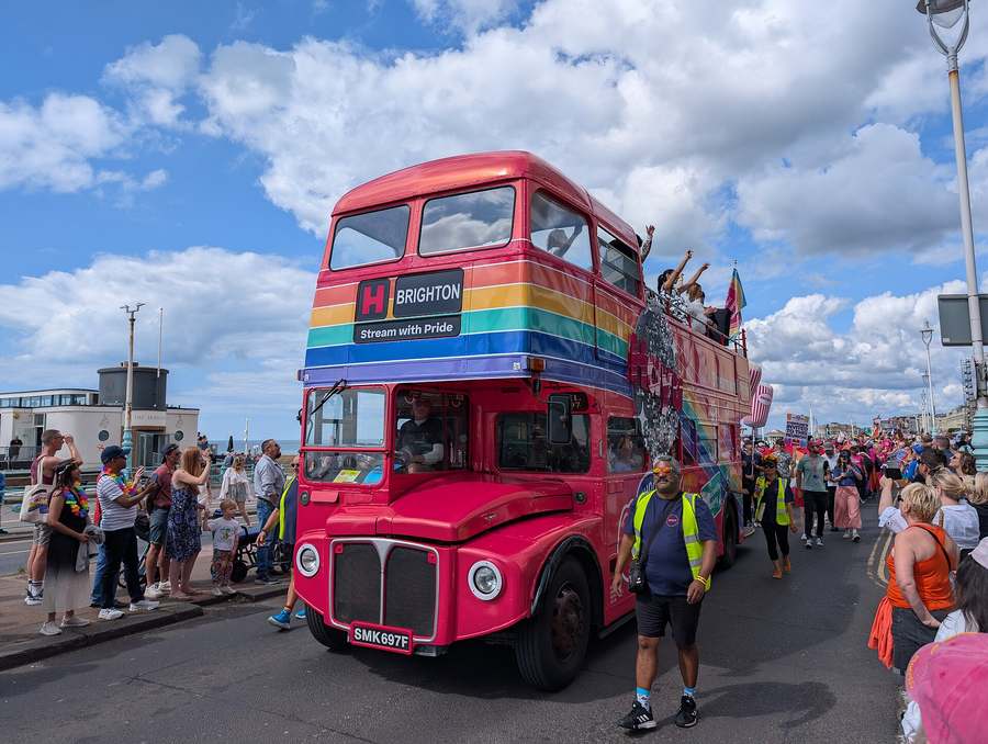 A rainbow painted double decker bus leads Pride parade-goers along the Brighton seafront under a blue sky