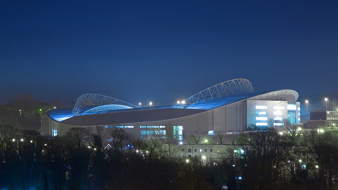 The Amex Stadium lit up at night with its curved roof glowing blue