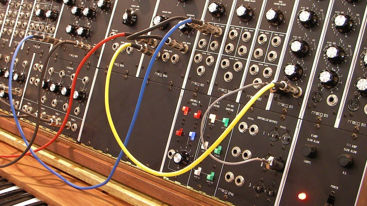 A close view of a modular synthesizer with coloured patch cables looping across black control panels, knobs and sockets