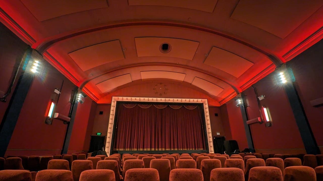 The empty auditorium of a historic cinema, with rows of red velvet seats facing a curtained screen under a curved ceiling lit in red