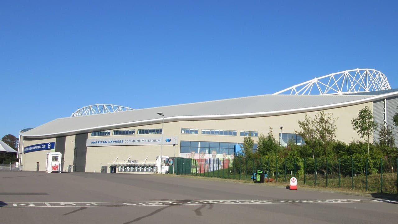 The outside of the American Express Community Stadium at Falmer under a clear blue sky, with its curved grey roof and white steel arches, and an empty car park in front