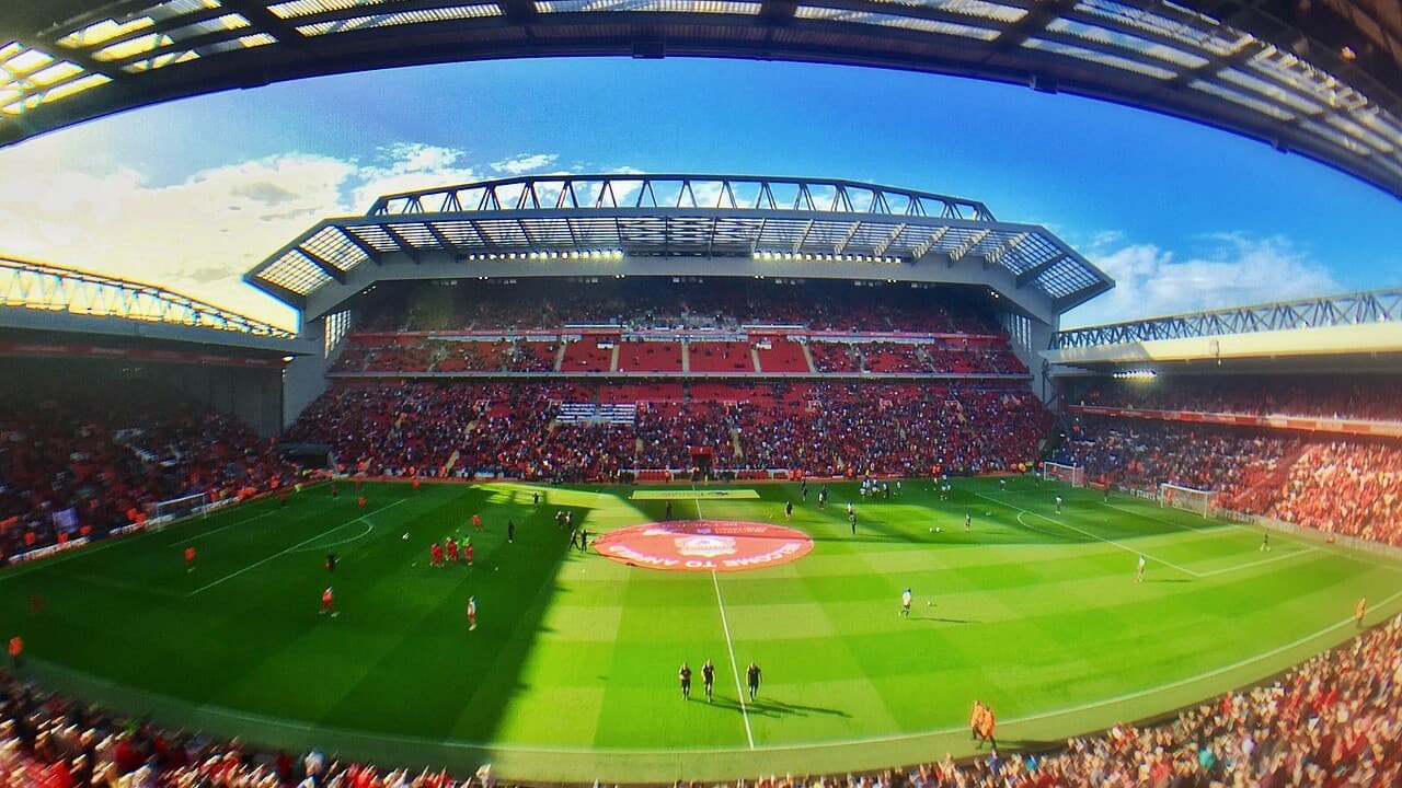 Anfield stadium before a match, with the Kop, the Main Stand and the Anfield Road Stand around the pitch under a blue sky
