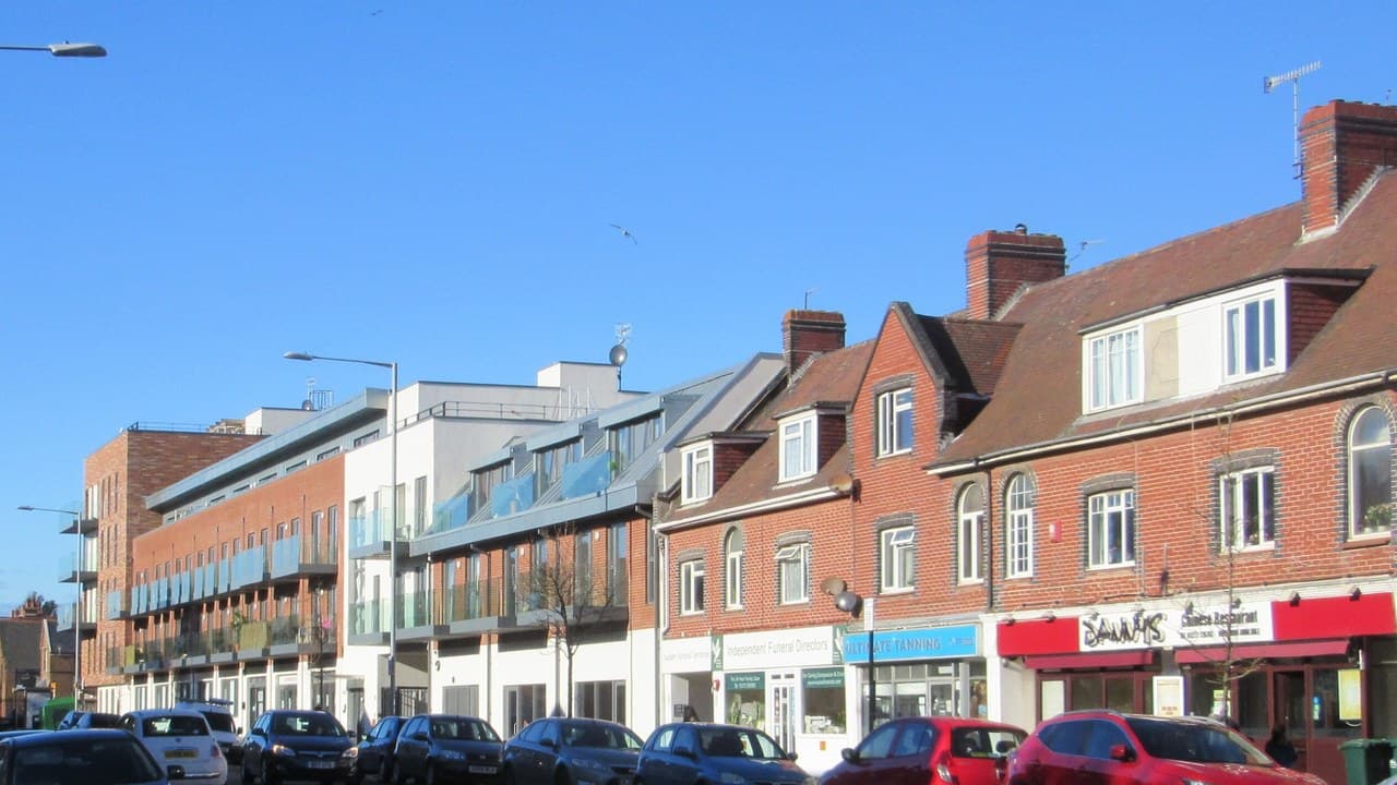 Portland Road in Hove on a sunny day, with shops under red brick flats, newer blocks of flats beyond and parked cars along the kerb