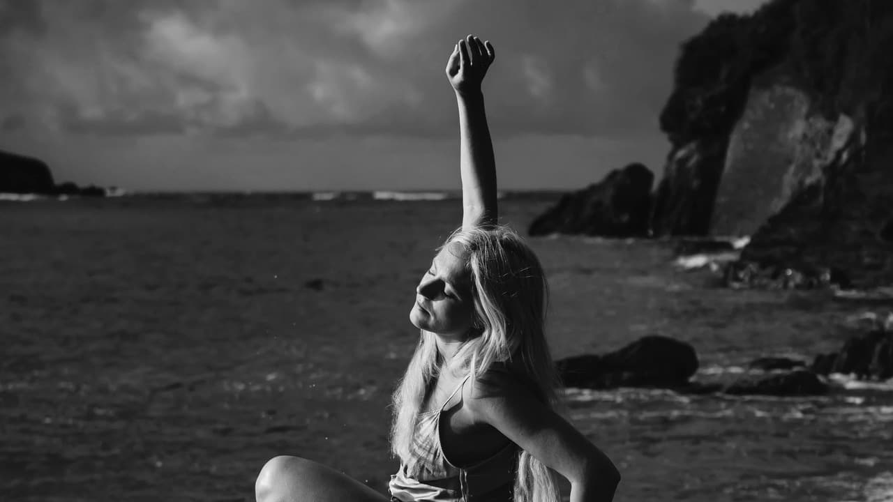 Black and white photograph of singer Tallulah Rendall sitting by the sea with one arm raised and her face tilted towards the light