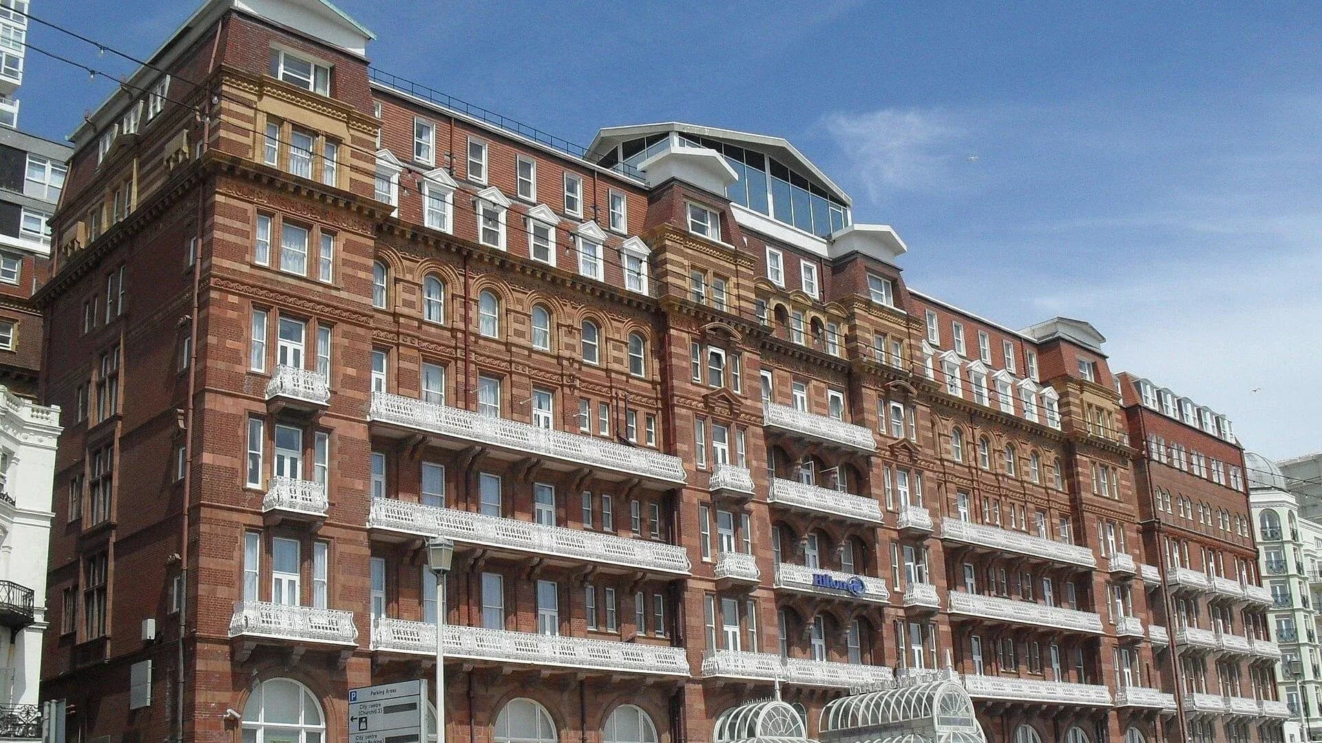 The red brick Hilton Brighton Metropole hotel on Kings Road, Brighton, with white balconies running along its front under a blue sky