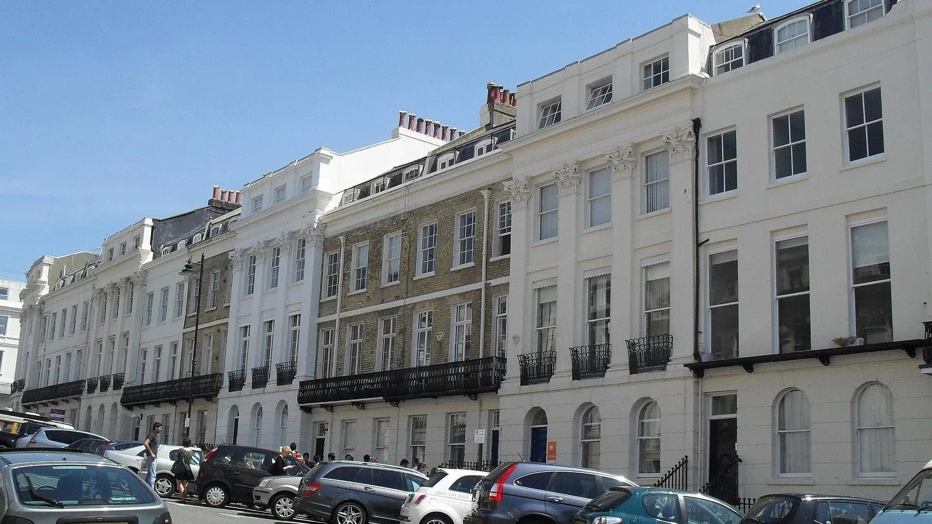 A long terrace of white and cream stucco houses with black iron balconies on Portland Place in Kemptown, Brighton, under a clear blue sky