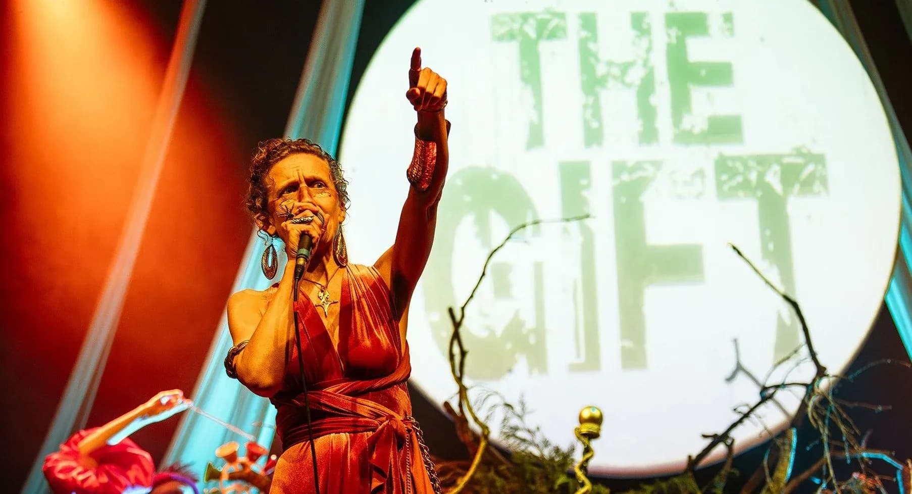 Mella Faye on stage at The Old Market in Hove, pointing upwards and holding a microphone in front of a large circular projection reading The Gift