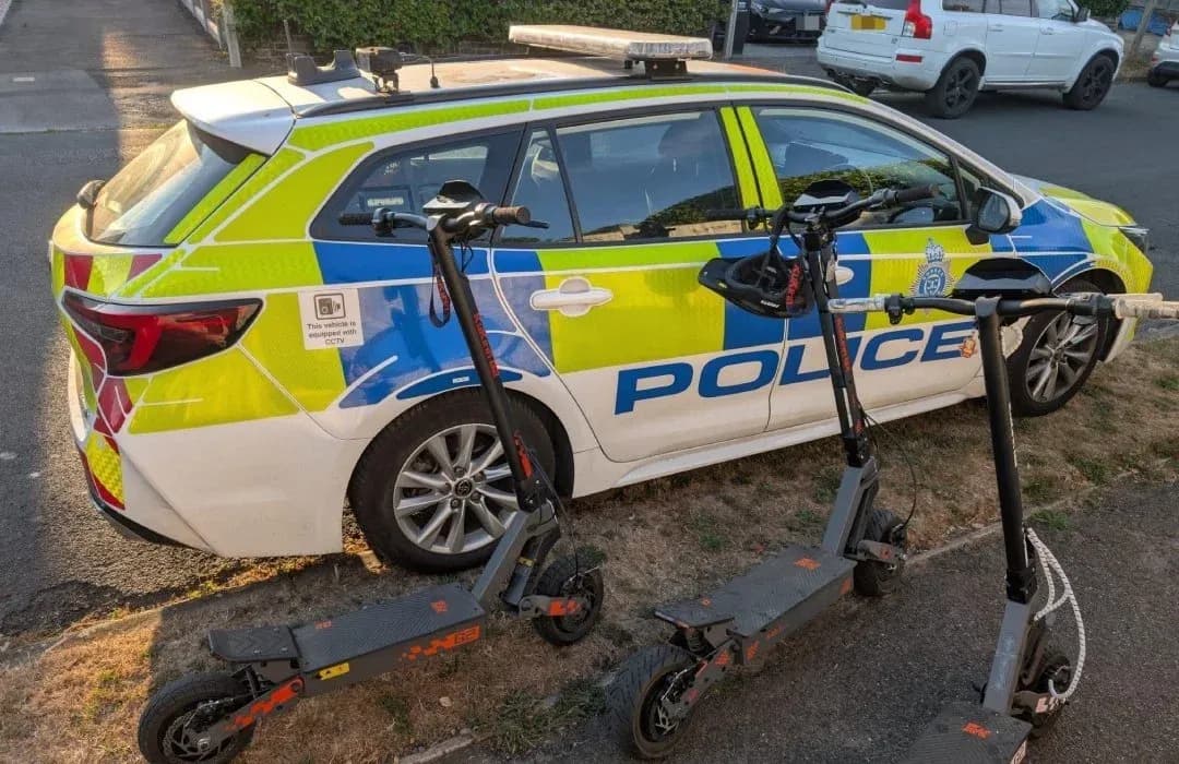 A Sussex Police estate car parked on a grass verge with three black and grey e-scooters standing in front of it