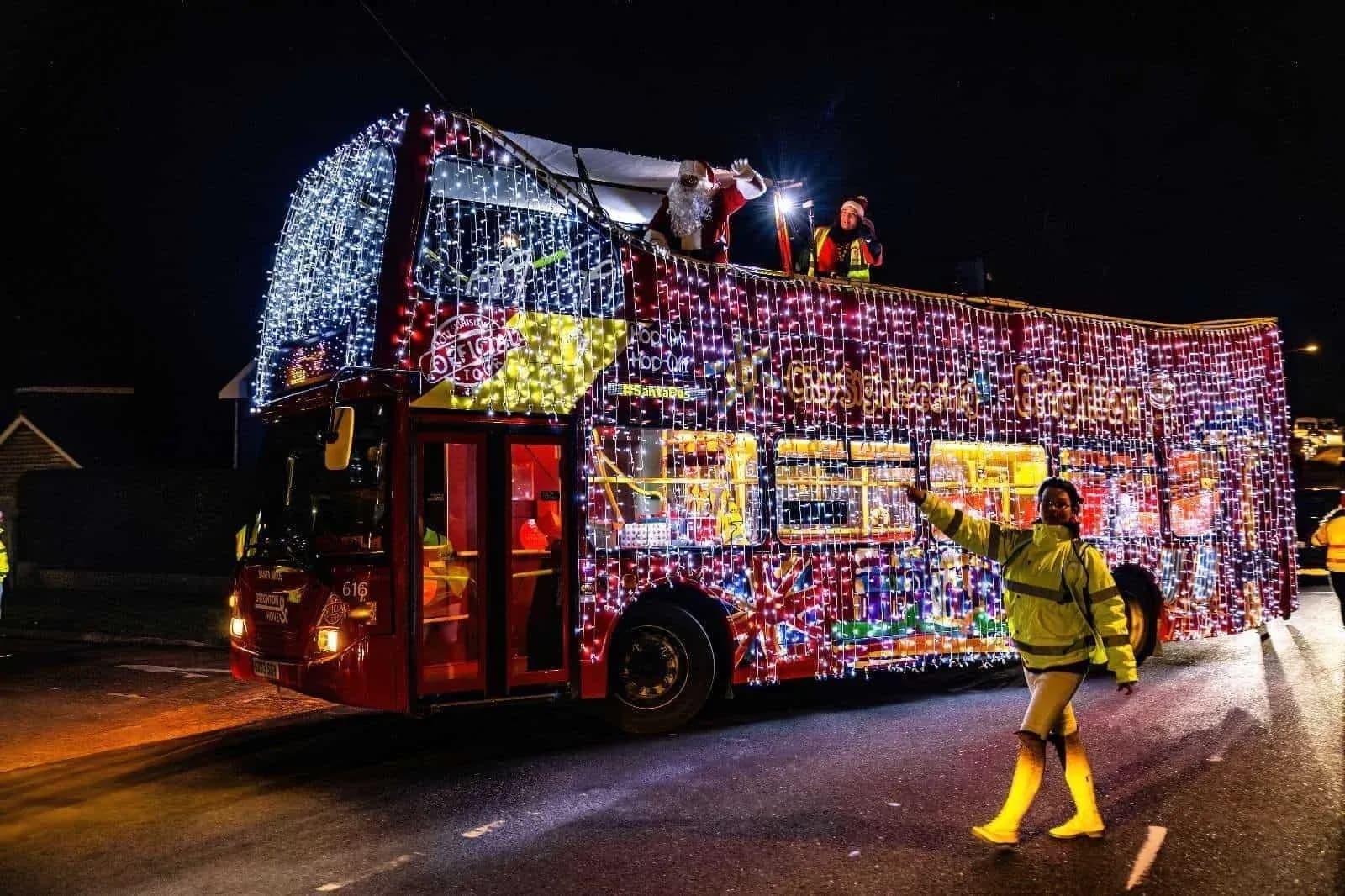The Santa Bus, a double decker covered in white and coloured fairy lights, driving through a street in Brighton and Hove at night with Santa waving from the top deck and a steward in a yellow jacket pointing the way