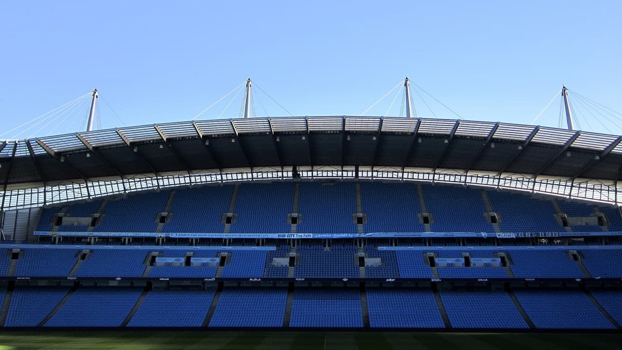 Etihad Stadium in Manchester seen from inside the stand on a clear day, with blue seats and the roof arches above the pitch